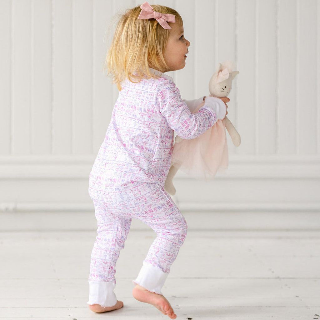 Side view of a child wearing a pink ballet zip onesie, holding a bunny toy against a white paneled background