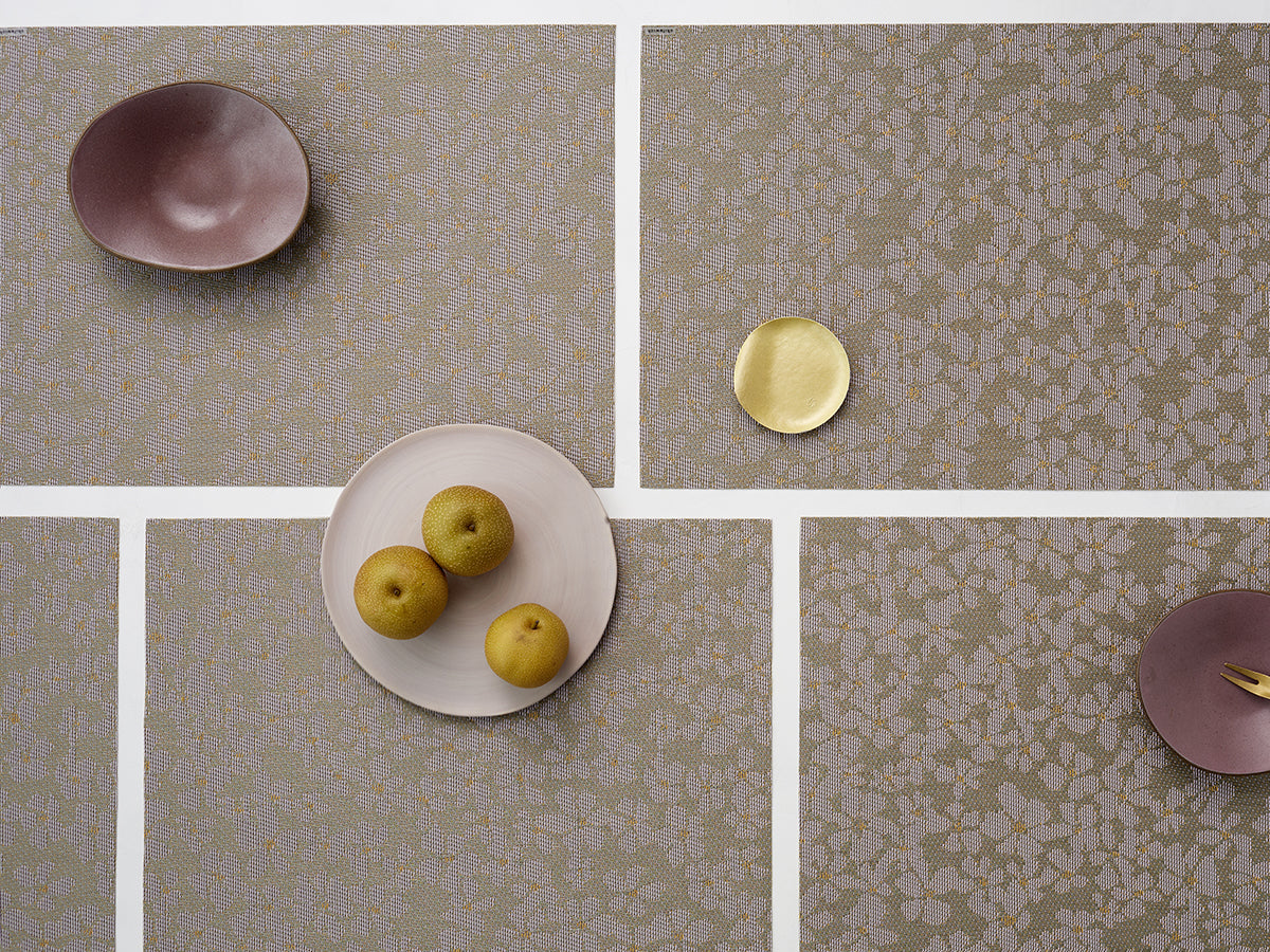 Overhead view of Flora placemat in mica showcasing textured floral weave and color accents on a white grid table with ceramic bowls