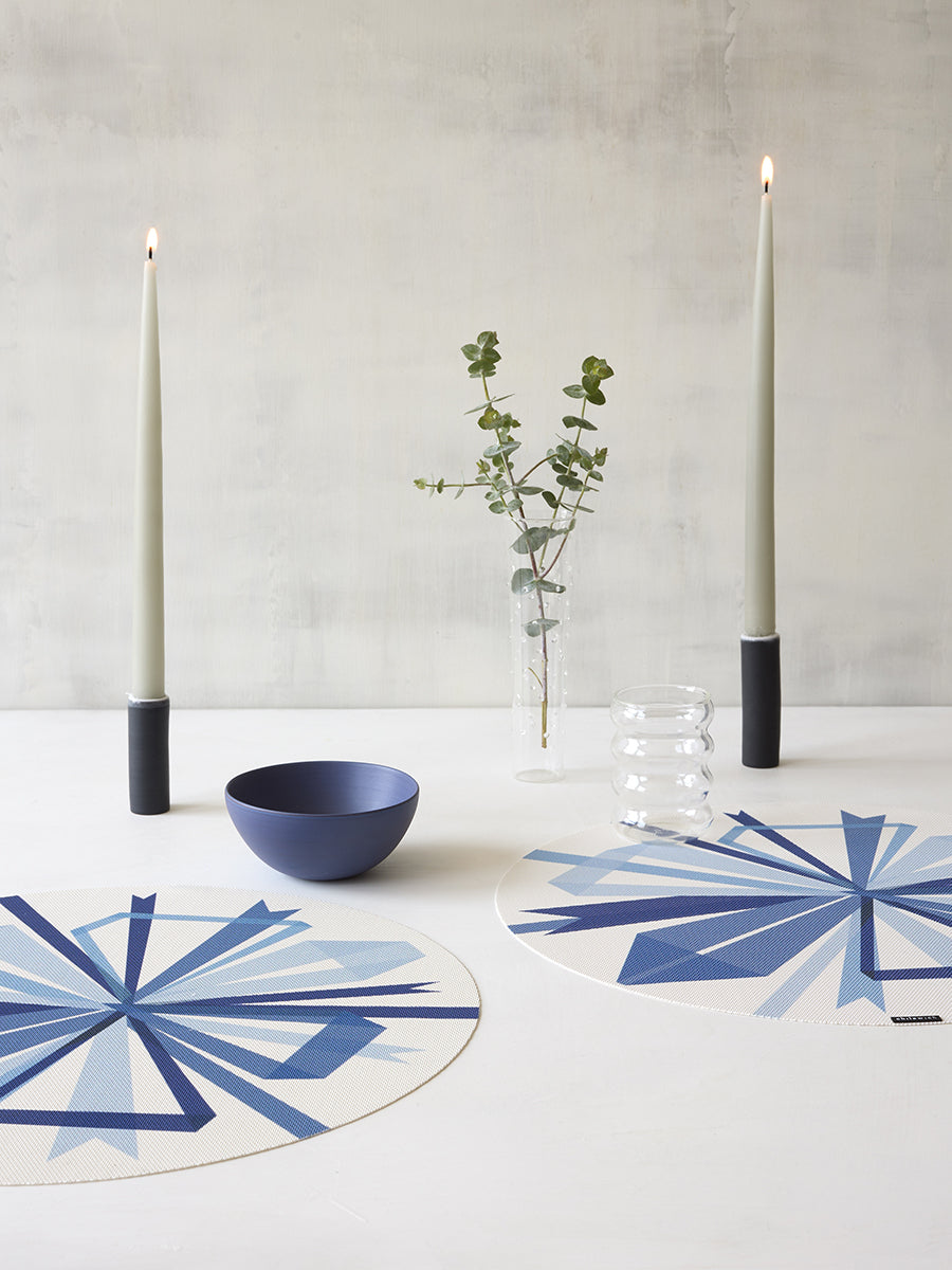 Flat lay of ribbon placemat in Frost showcasing blue geometric starburst print and woven texture on a white table with candles and bowl