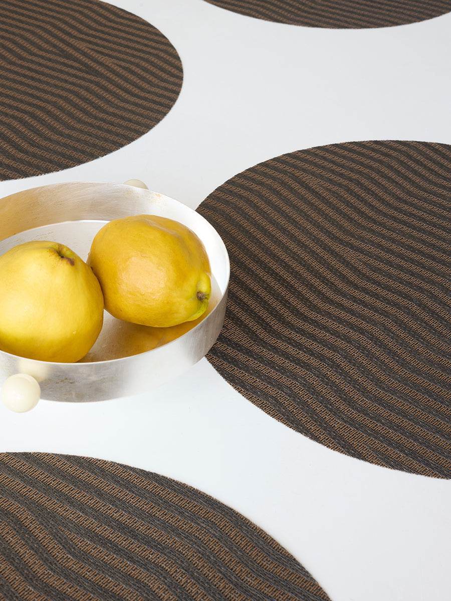 Angled close-up of Dune Round placemat in Raisin showing wavy woven texture on white table with silver bowl and yellow fruit