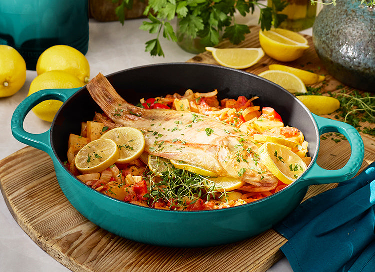 Angled overhead of 11-inch Riviera Signature Everyday Pan showing teal enamel finish, two handles, with cooked fish and lemon on a wooden board