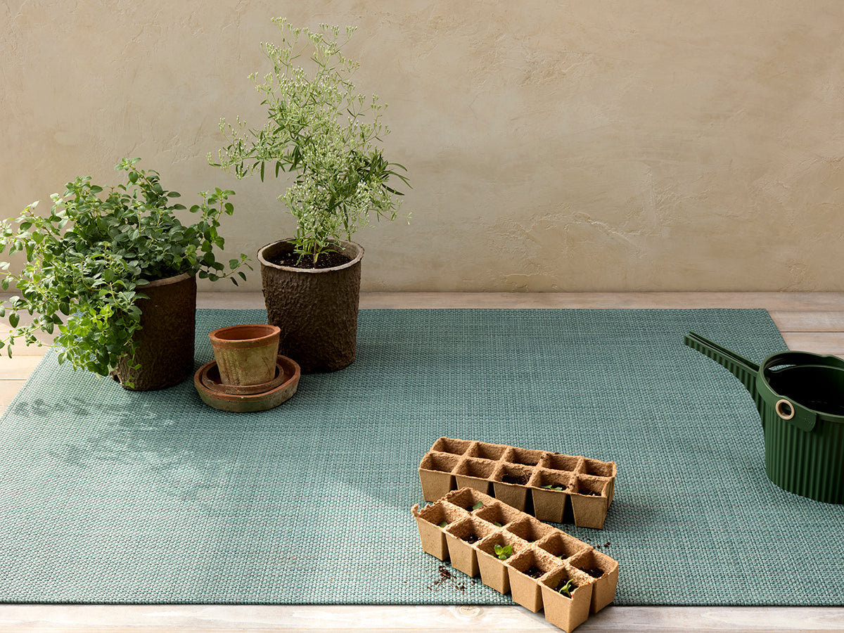 Angled top-down view of Basketweave rectangular placemat in celadon showcasing woven texture with potted plants, seedling trays, and a watering can on a light wood floor