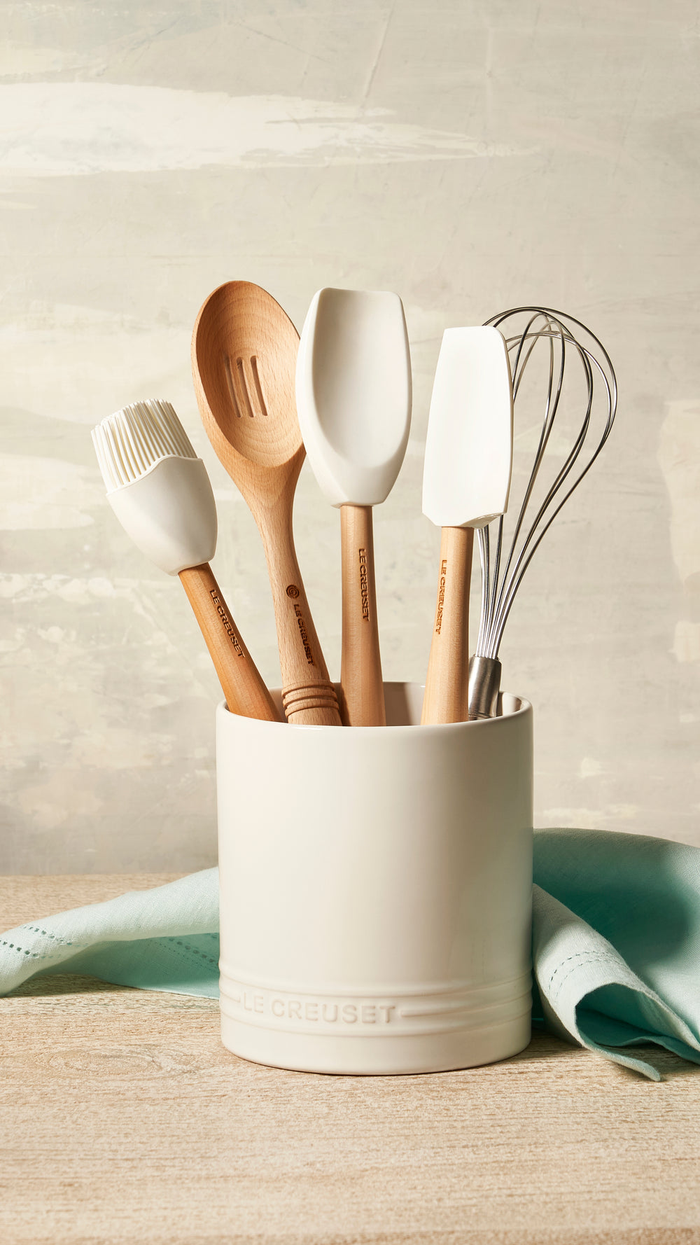 White silicone spatula spoon standing in a cream utensil crock with a wooden spoon, brush, and whisk on a countertop