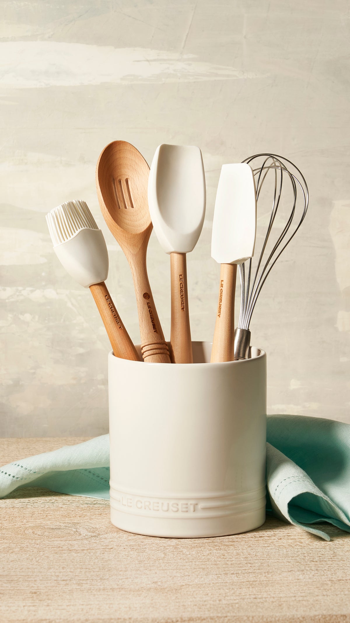 White silicone spatula spoon standing in a cream utensil crock with a wooden spoon, brush, and whisk on a countertop
