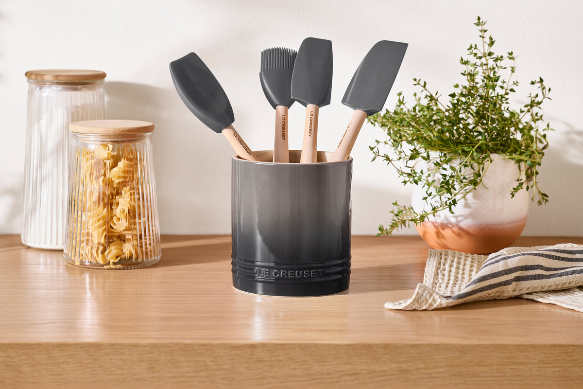 Front view of Signature Utensil Crock in oyster holding silicone utensils, showing straight sides and glossy glaze on a wooden countertop with jars and a potted plant