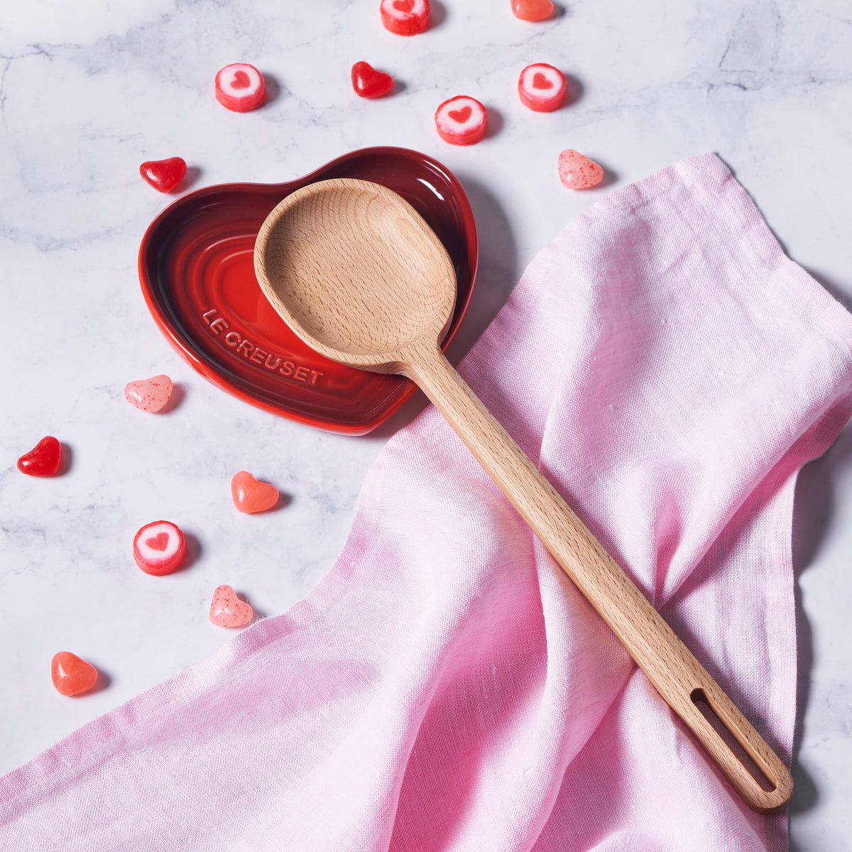 Top-down view of Signature wood spoon showcasing beech wood bowl and long handle resting on pink linen and a red heart dish