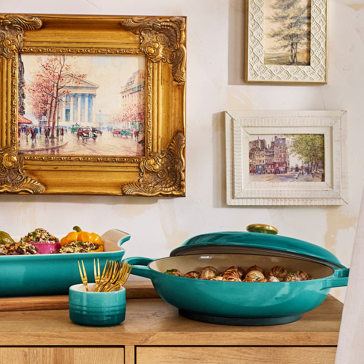 Riviera straight-wall ramekin holding gold forks on wooden buffet beside matching covered casserole with framed art behind
