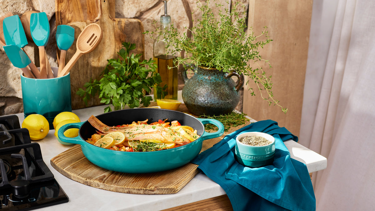 Riviera spatulas in a utensil crock beside a matching turquoise Le Creuset skillet with cooked fish and lemon on a kitchen counter