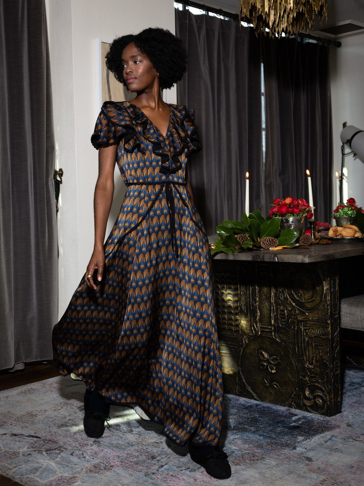 Three quarter view of Keepsake Dress in Midnight on model showing ruffled V-neck, fitted waist, and blue and gold geometric print beside a candlelit dining table
