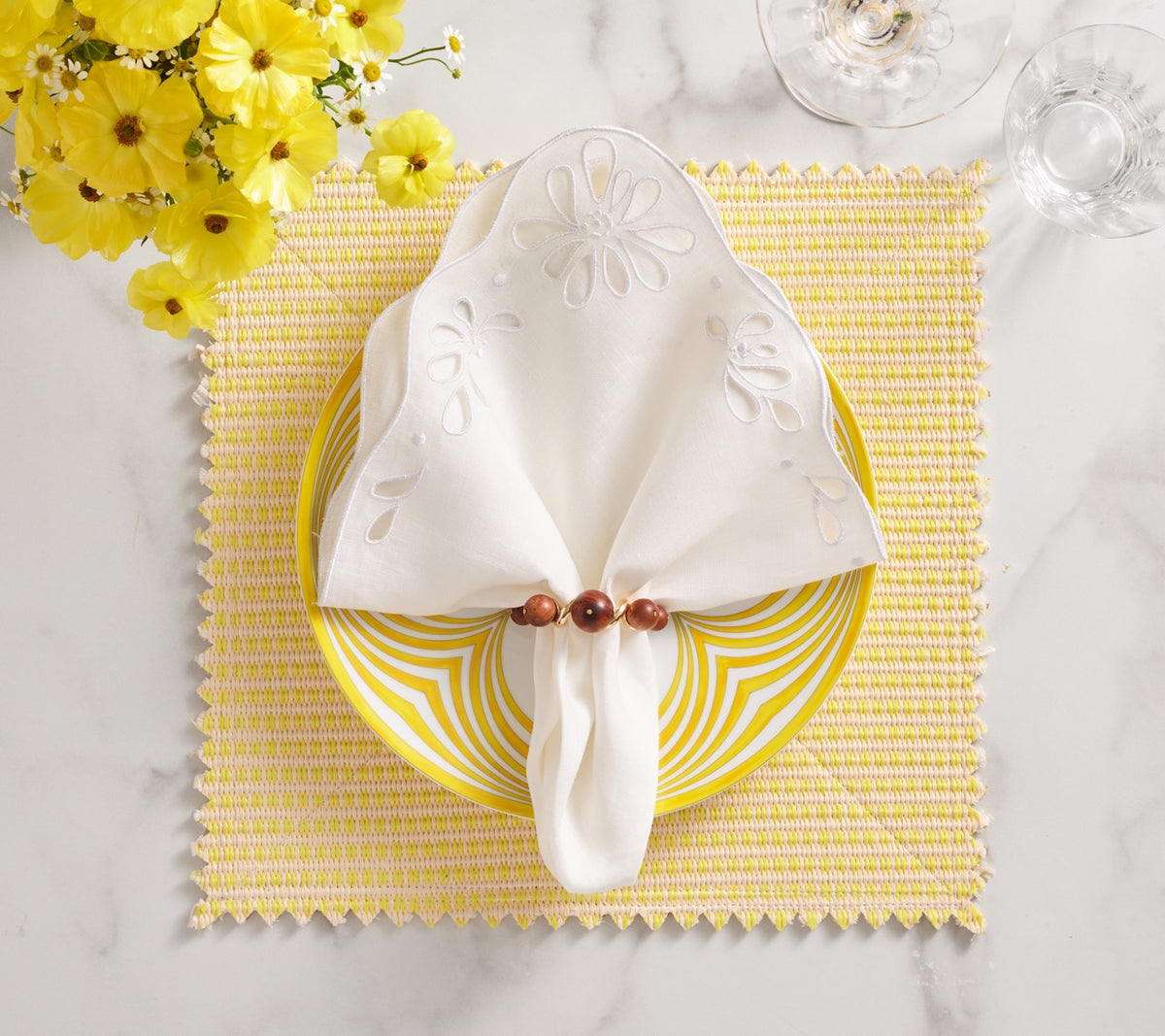Top view of Bloom Napkin in white showing eyelet embroidery, folded in a wooden bead ring on a yellow plate and placemat