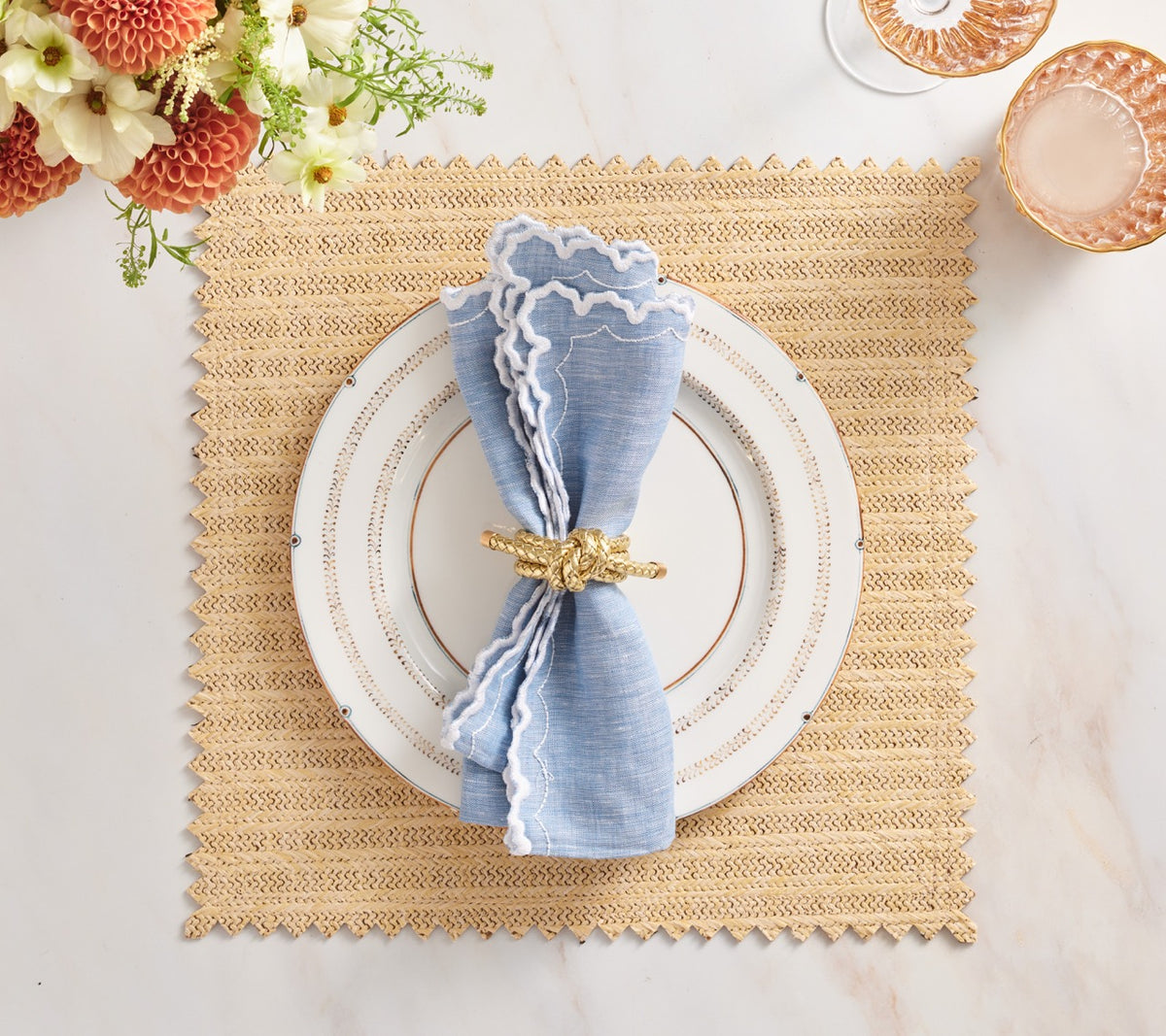 Top view of Arches napkin, blue with white scalloped border, folded with gold knot ring on white plate over woven placemat