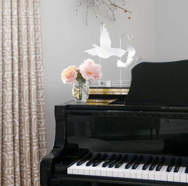 Two acrylic dove ornaments displayed on top of a black grand piano with a vase of pink peonies and patterned curtains