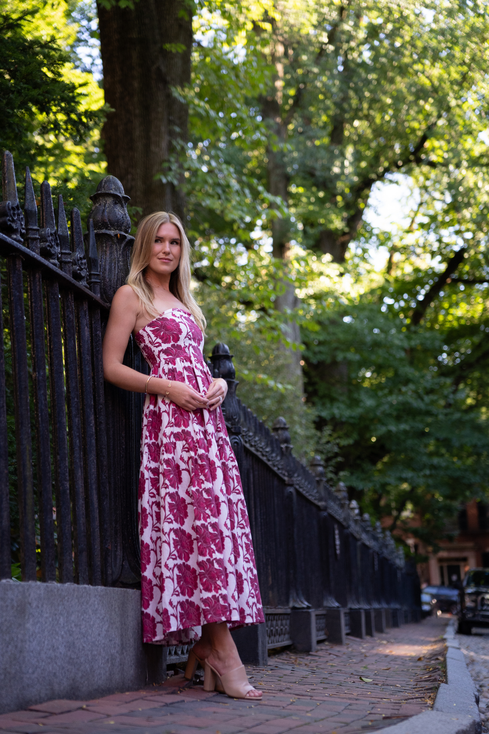 Full-length view of Barbie Dress in Dahlia on model, strapless floral bodice and mid-calf skirt, iron fence and trees