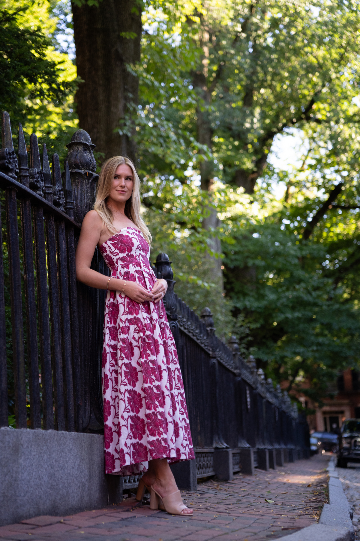 Full-length view of Barbie Dress in Dahlia on model, strapless floral bodice and mid-calf skirt, iron fence and trees