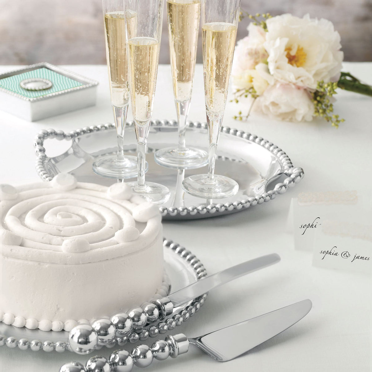 Angled close-up of Pearled Large Oval Platter showing beaded pearled rim with champagne flutes, a white frosted cake, and pale flowers on a white table