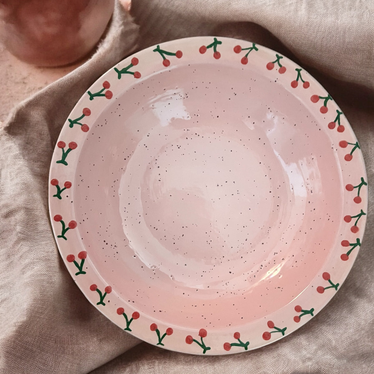 Top view of hand-painted cherry bowl showcasing pink speckled glaze and painted cherry rim on beige linen background