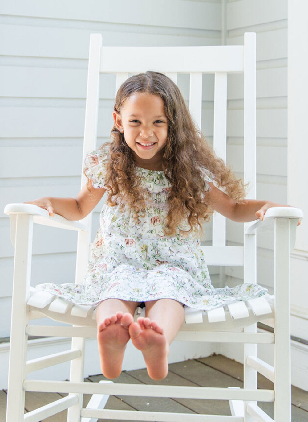 Front view of The Mimi Dress in Hunter Pace print on a child showcasing smocked bodice and ruffle sleeves on a white rocking chair