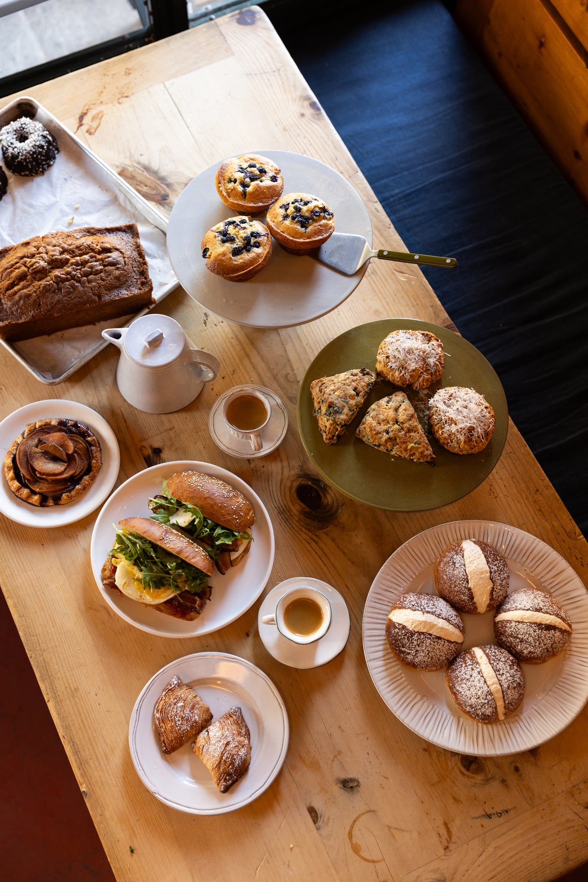 White pedestal side plate holding three blueberry muffins on a wooden table with pastries, sandwiches, and espresso in a casual cafe setting