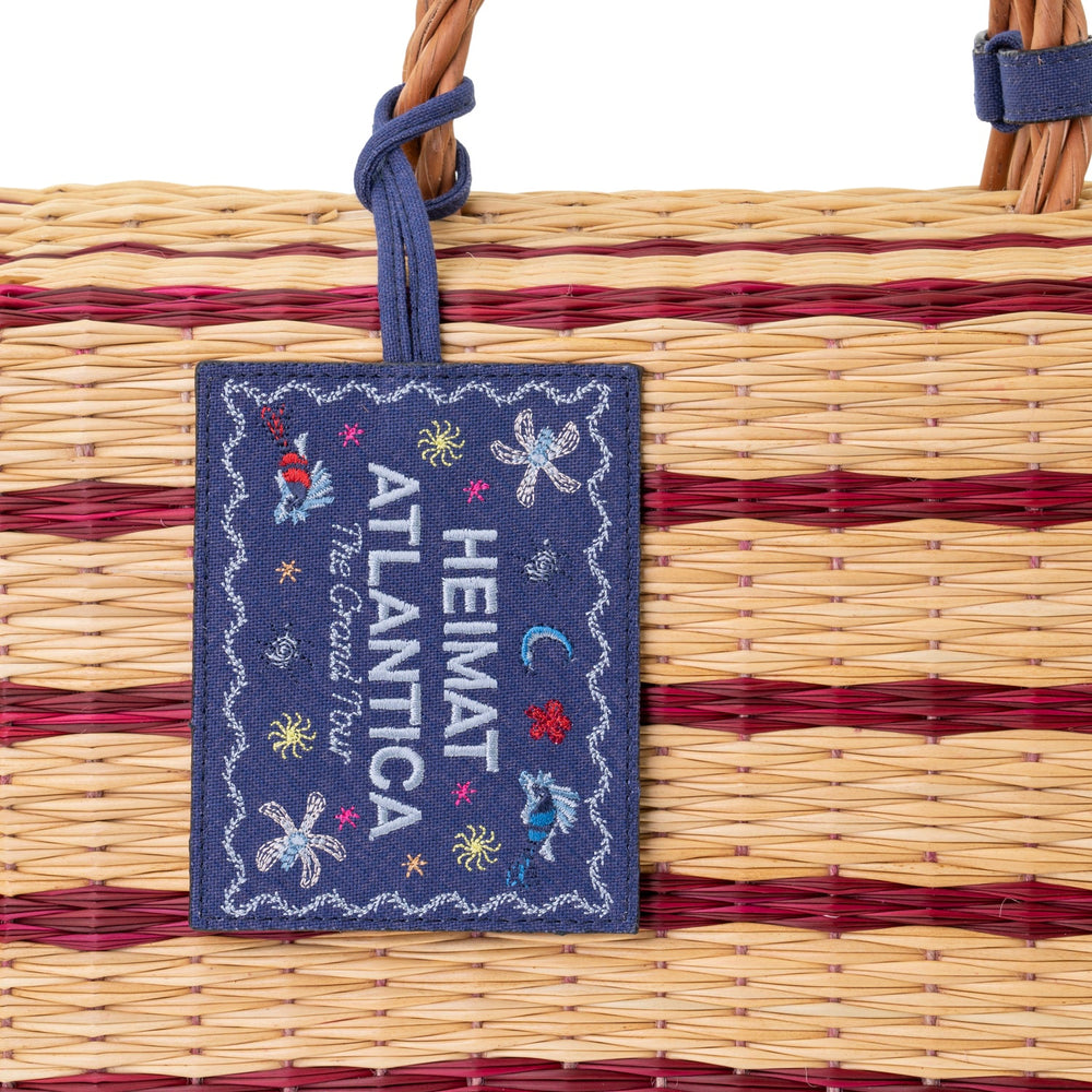 Close-up of The Grand Tour Collection beach basket showing burgundy-striped handwoven reed and navy embroidered label reading HEIMAT ATLANTICA