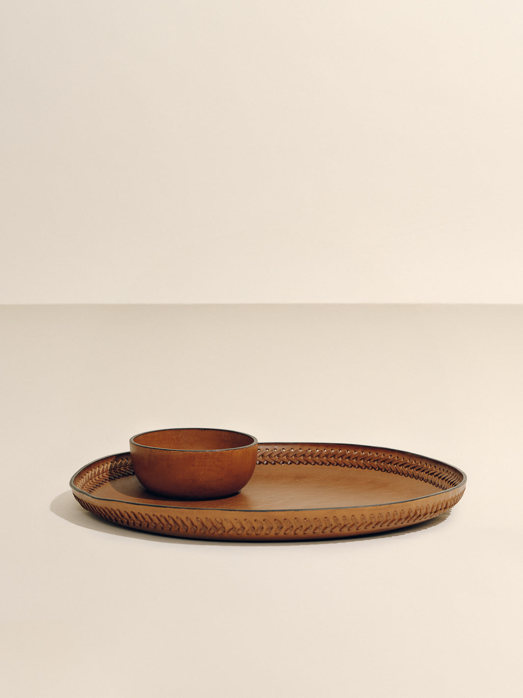 Angled view of Oversized Tray in cognac leather showcasing handwoven lace trim with small bowl on neutral beige background