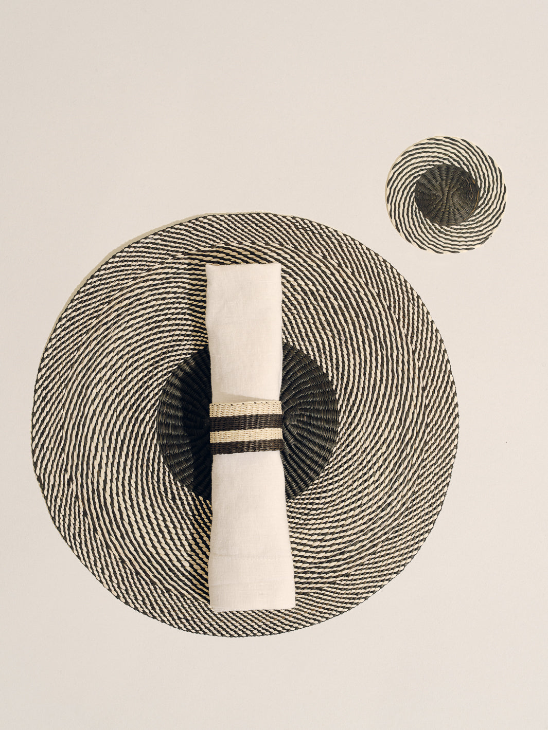 Top-down view of woven coaster in natural black showing spiral two-tone weave with rolled white napkin and striped napkin ring