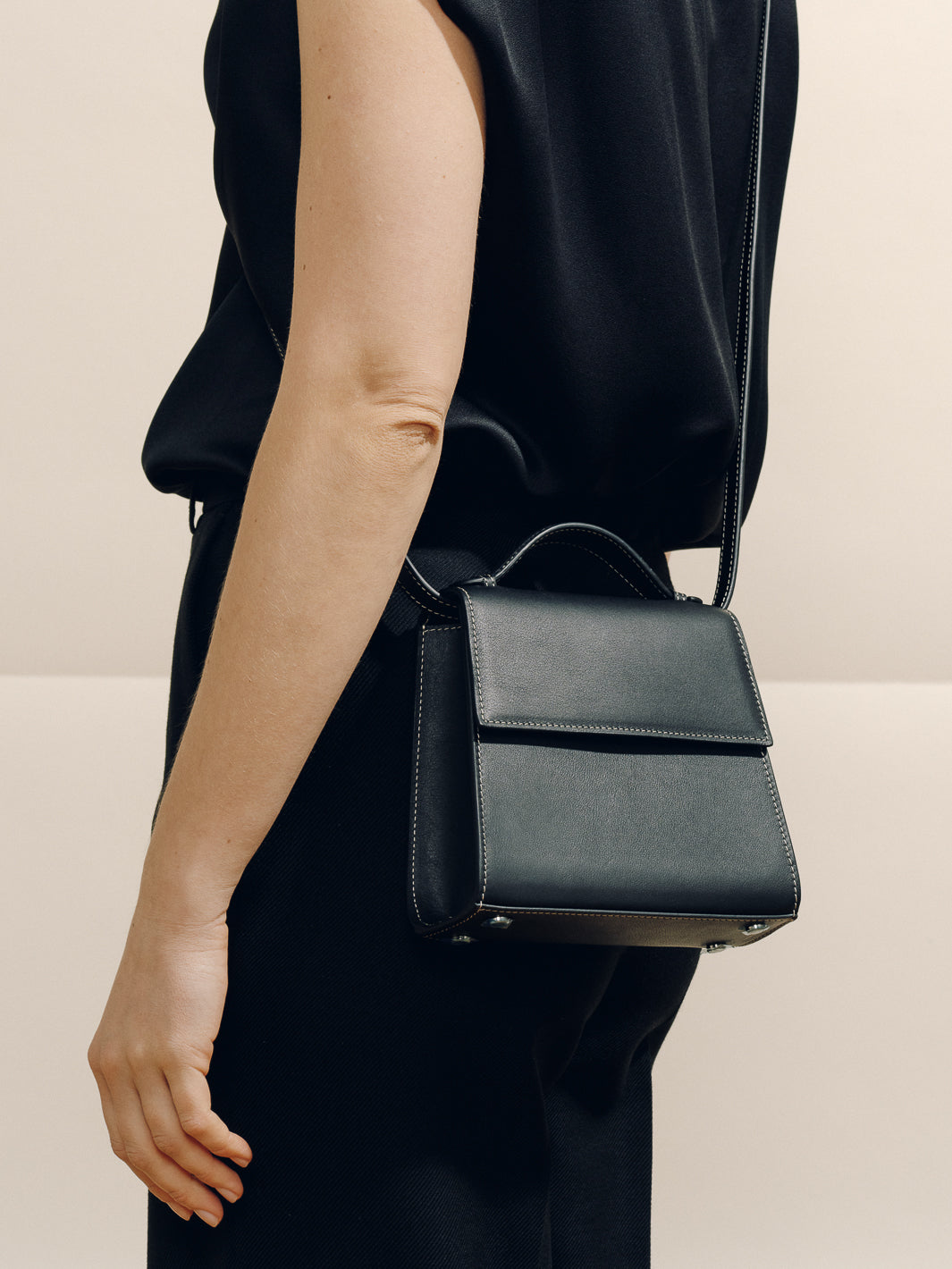 Side view of The Small Top Handle bag in black nappa showcasing top handle, structured box shape and contrast stitching against a neutral beige background