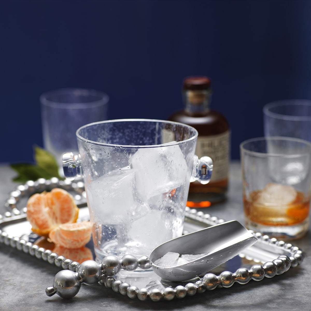 Beaded medium service tray with ice bucket, metal ice scoop, peeled tangerine and glasses on a gray tabletop