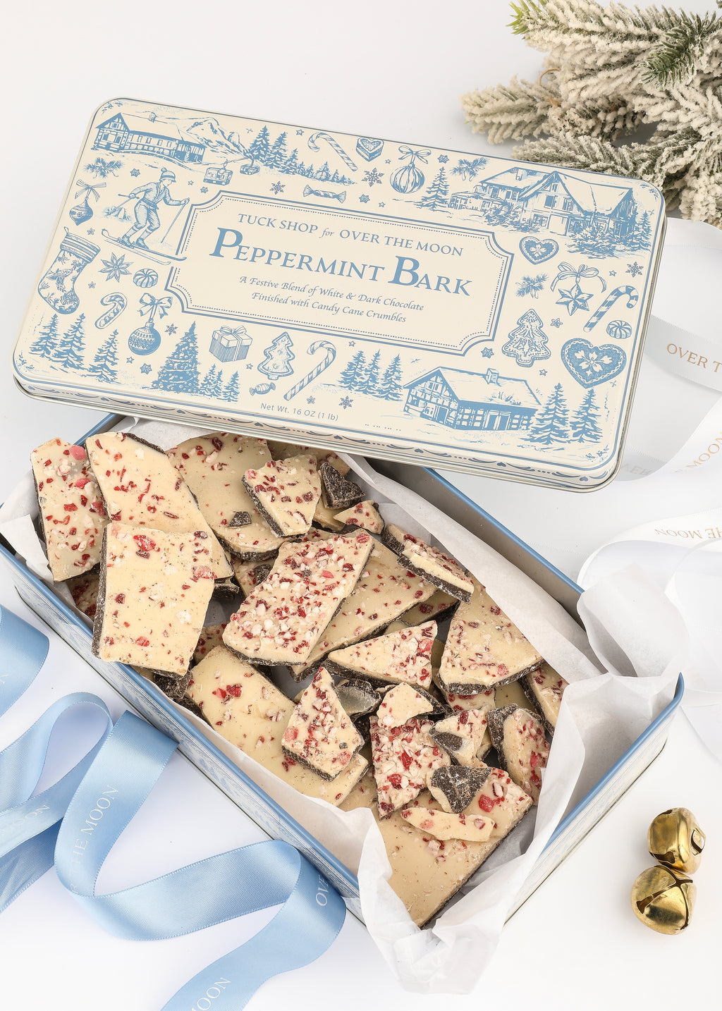 Top view of Over The Moon peppermint bark in a keepsake tin with peppermint-topped pieces, greenery, and ribbon