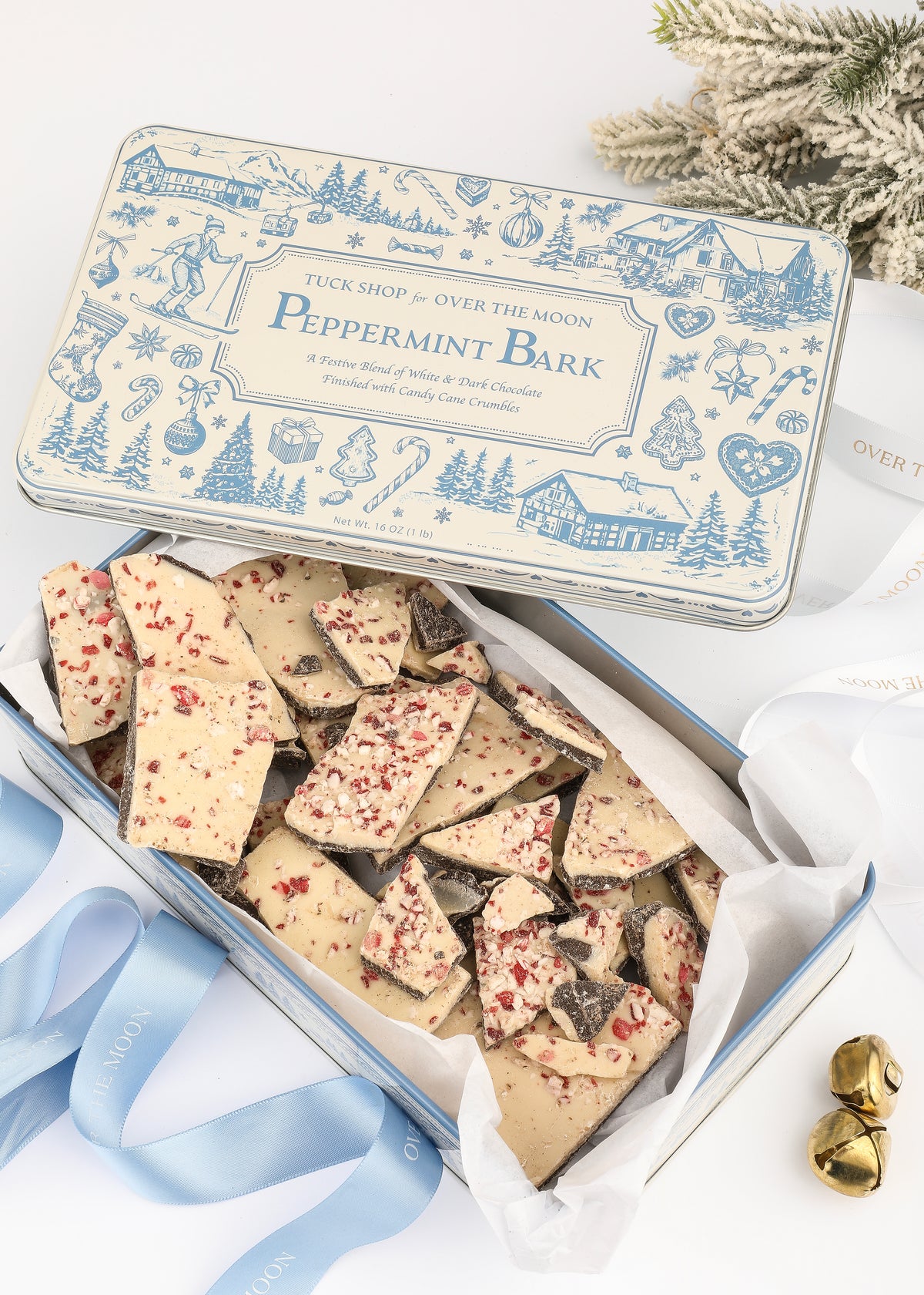 Top view of Over The Moon peppermint bark in a keepsake tin with peppermint-topped pieces, greenery, and ribbon
