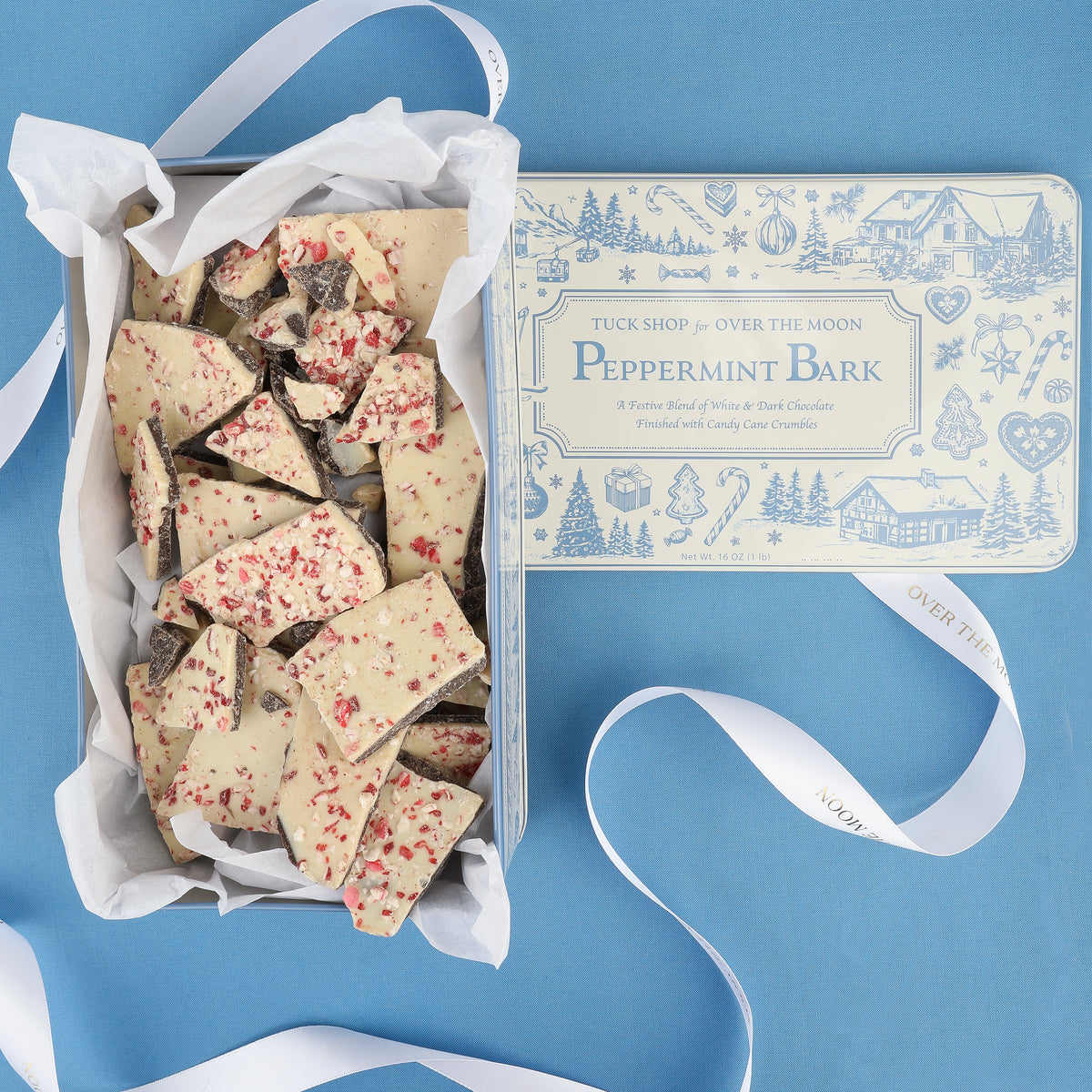 Overhead view of Over The Moon peppermint bark pieces with peppermint topping and keepsake tin on blue