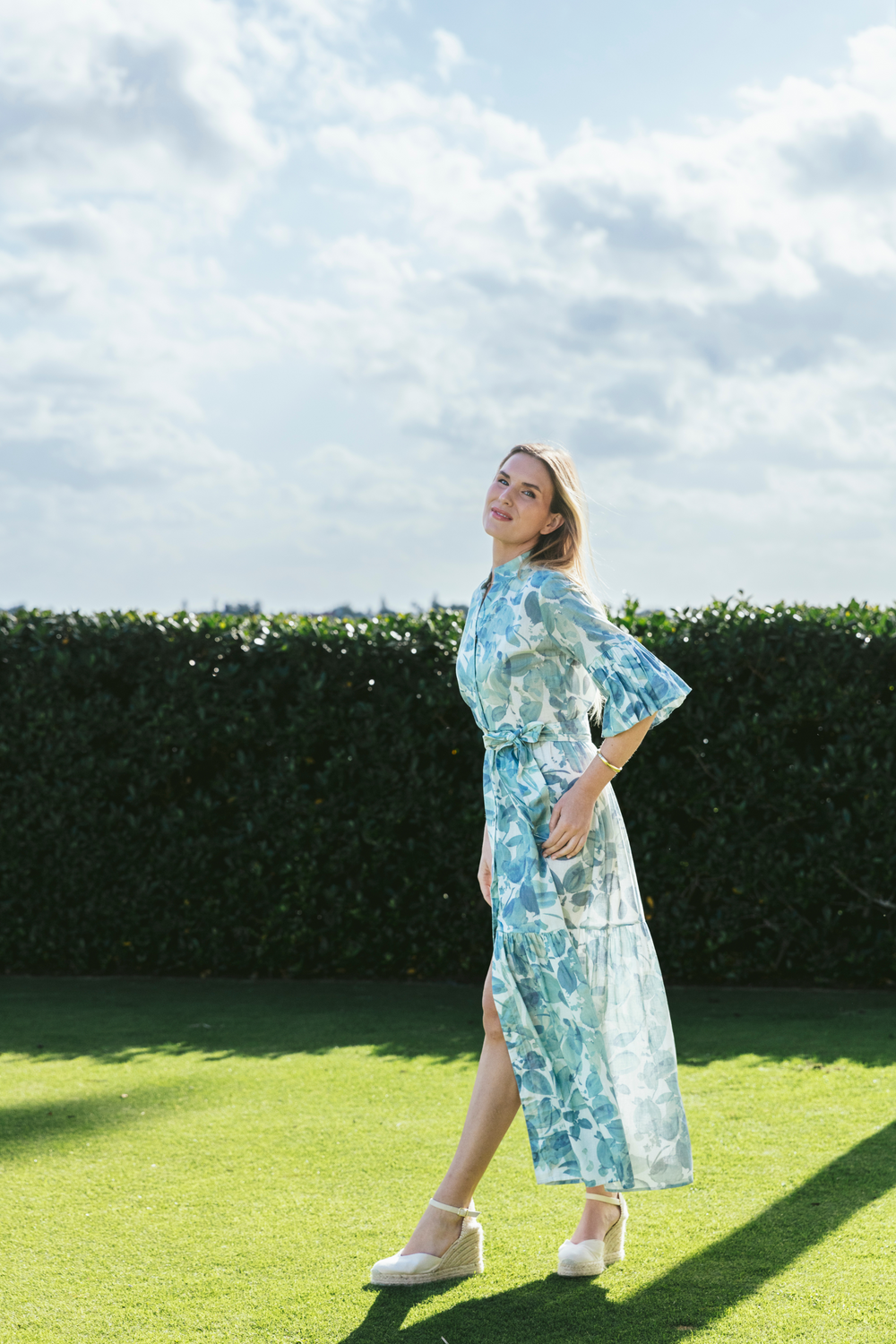 Three-quarter view of Joanna dress in blue-green floral linen showing bell sleeves, sash, tiered skirt and side slit on lawn