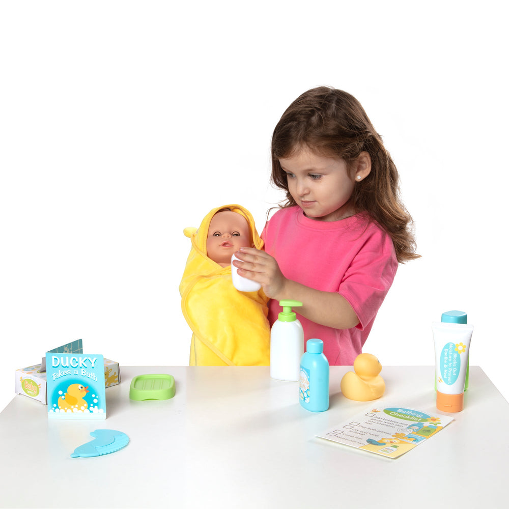 Front view of Mine-to-Love Changing & Bathtime Play Set showing a child holding a doll wrapped in a yellow hooded towel with bath bottles, rubber duck, and bath book on a white table
