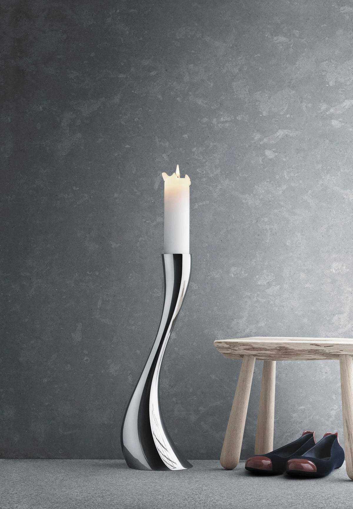 Front view of Cobra floor candleholder in mirror-polished stainless steel showing its sinuous curve with lit white candle beside a wooden stool on a gray textured background
