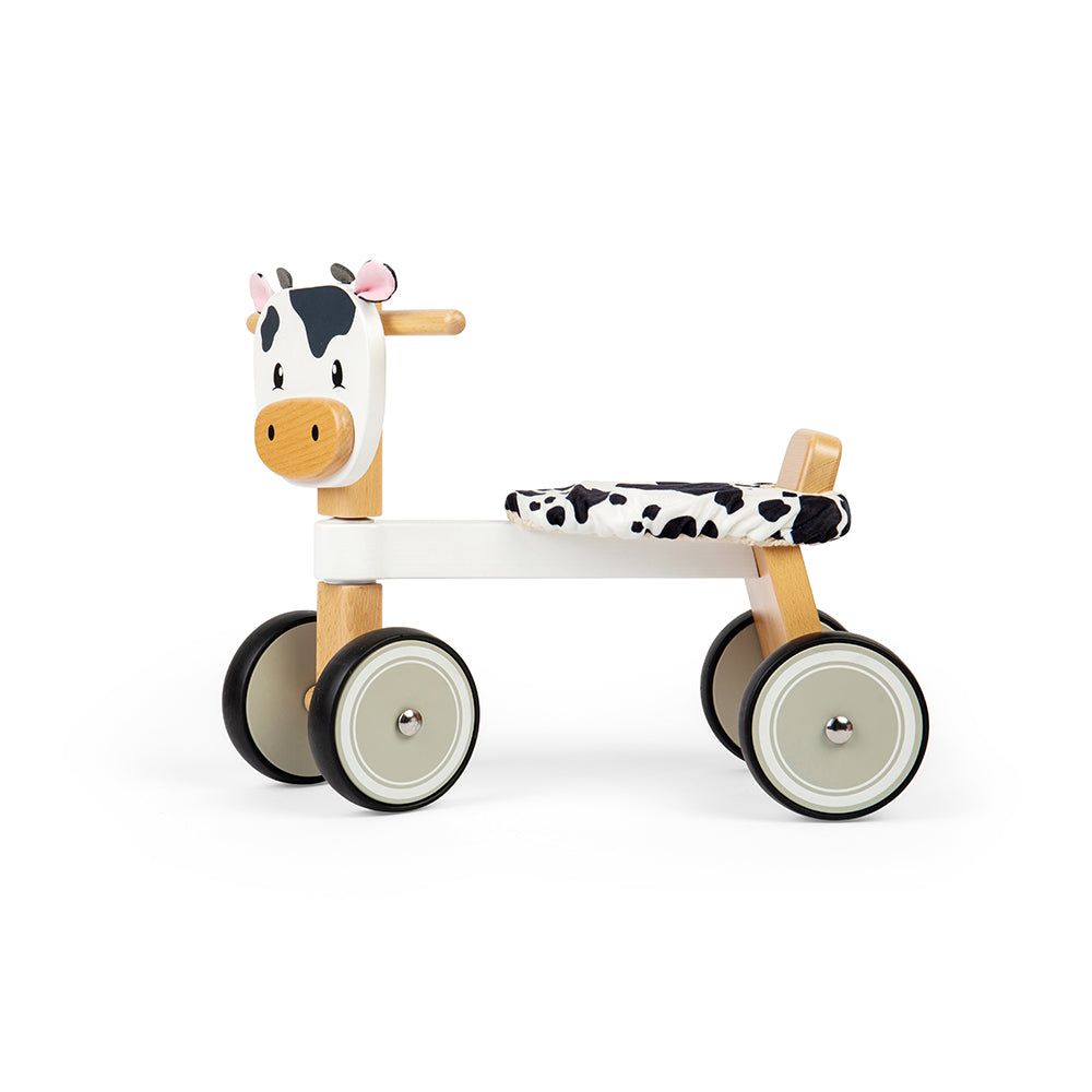 Side view of Ride On Cow showcasing four chunky wheels, wooden handlebars, and fluffy cow print seat with white background