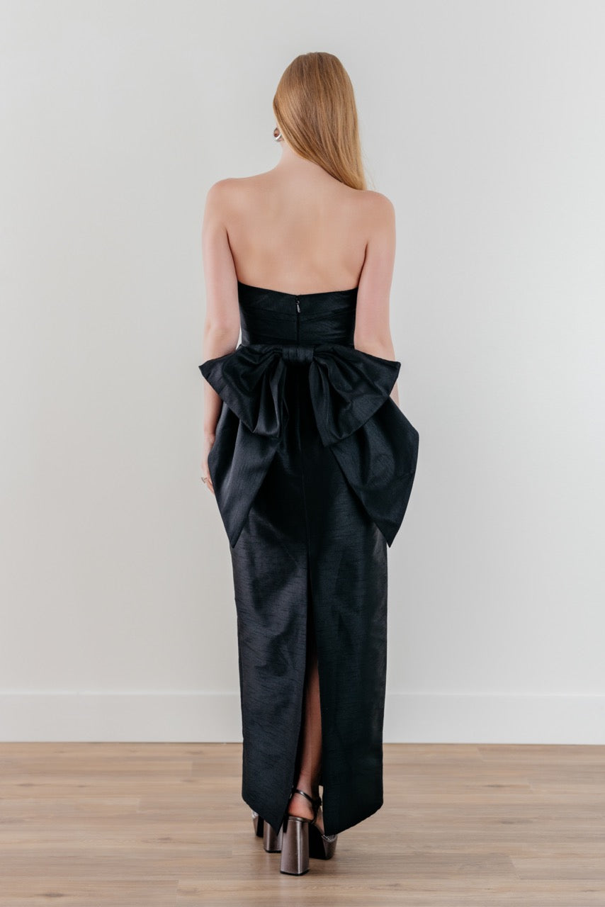 Rear view of Caladium dress in black showcasing strapless bodice, oversized detachable back bow and center back slit on white studio background