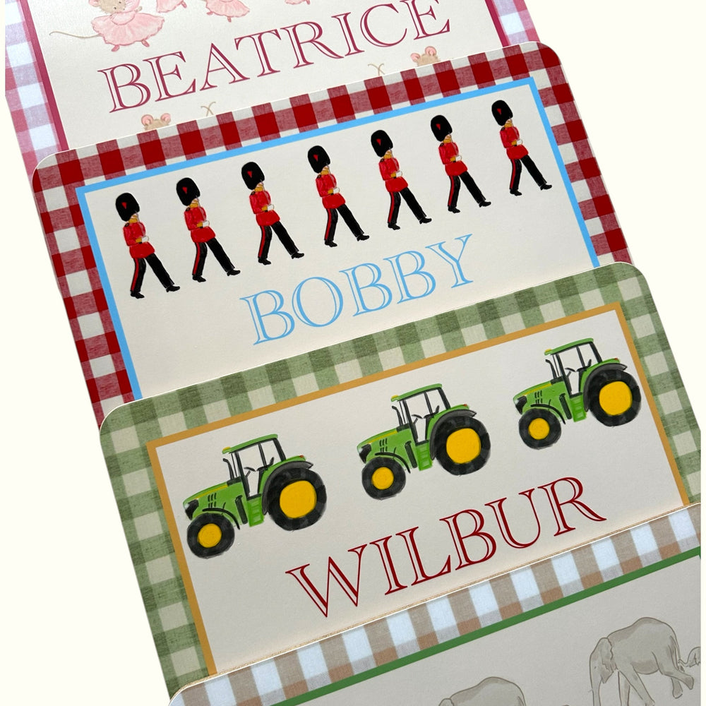 Angled view of three personalized table mats with gingham borders named Beatrice, Bobby, and Wilbur, each with illustrations