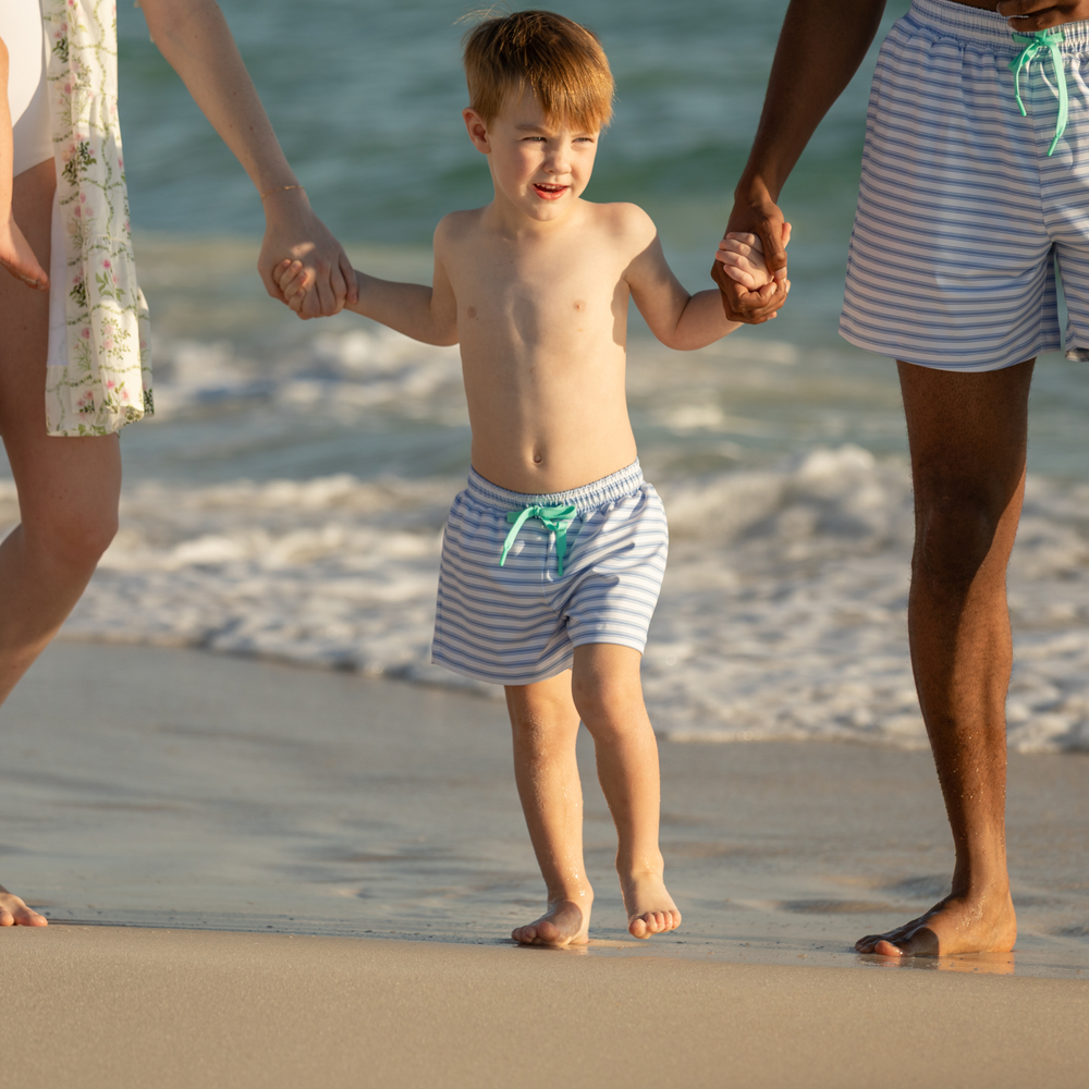 Young boy holding adults’ hands on a sandy beach wearing blue-and-white Bay Street Stripe swim trunks with green drawstring