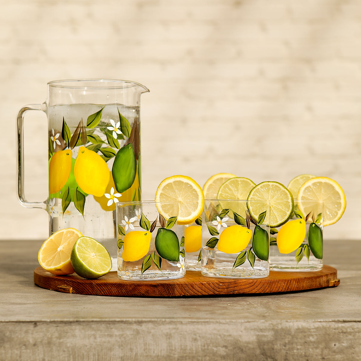 Front view of citrus jug with tumblers showing hand-painted lemon and lime motif on clear glass on wooden board