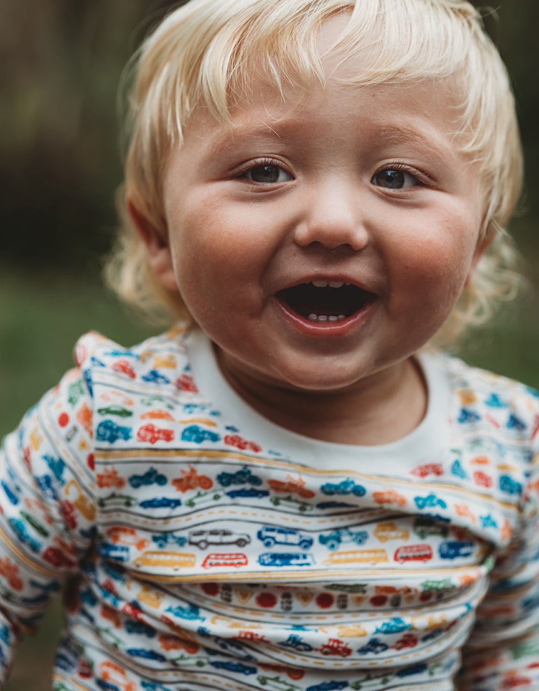 Front view of white cars and trucks two-piece pajamas showcasing a colorful vehicle print on a smiling child with a blurred background