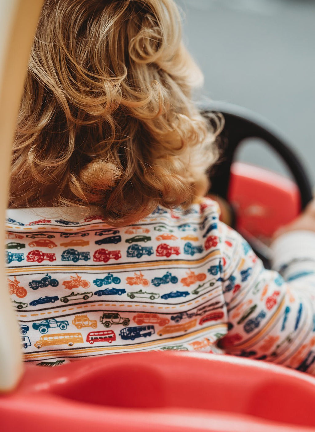 Child wearing cars and trucks pajamas while sitting in a red ride-on car