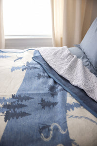Blue and cream blanket with ski and evergreen tree pattern, partially folded on bed with gray pillow.