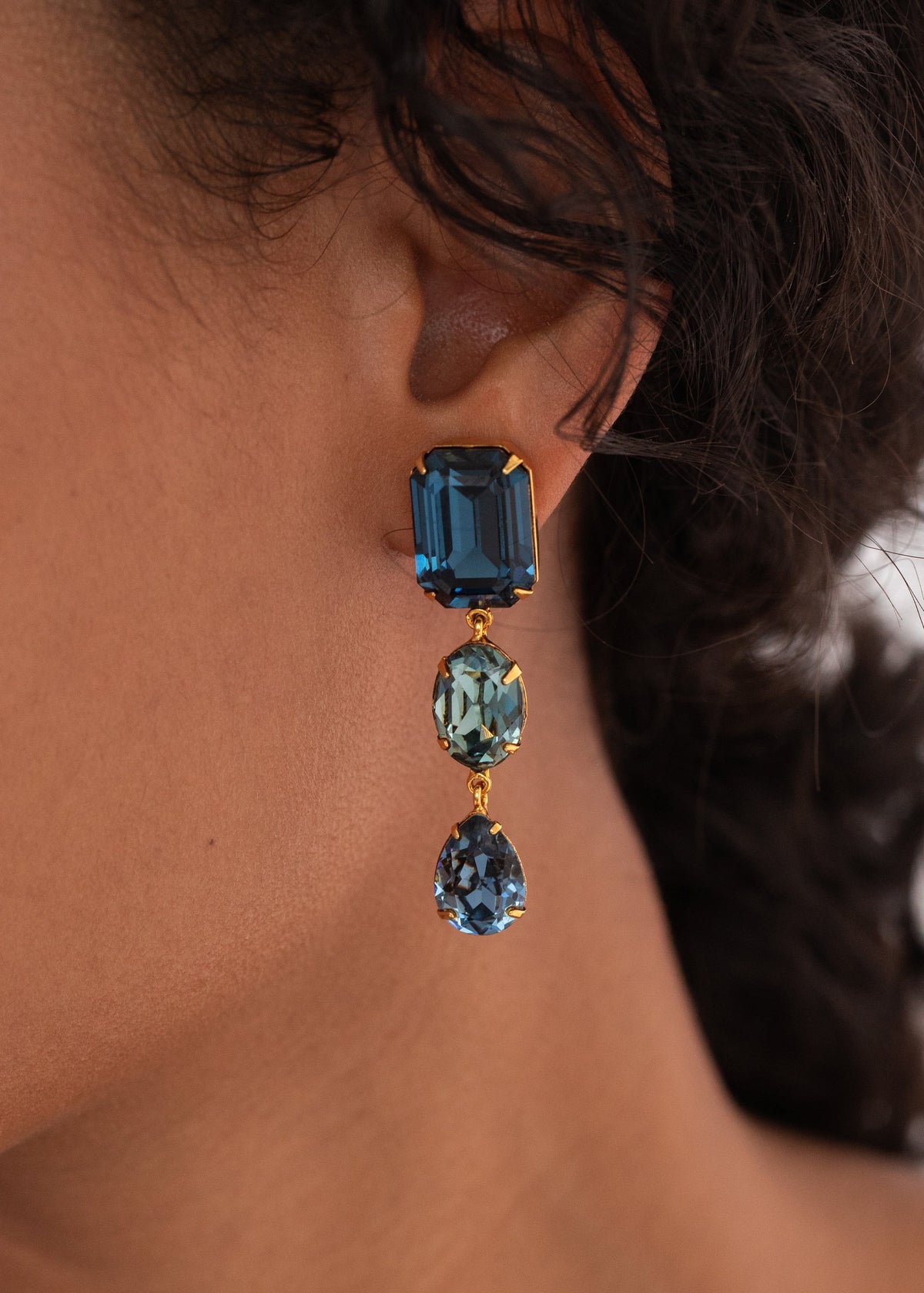 Close-up of Aviva Earrings in Sapphire showcasing three crystal cuts on a model’s ear with dark hair background