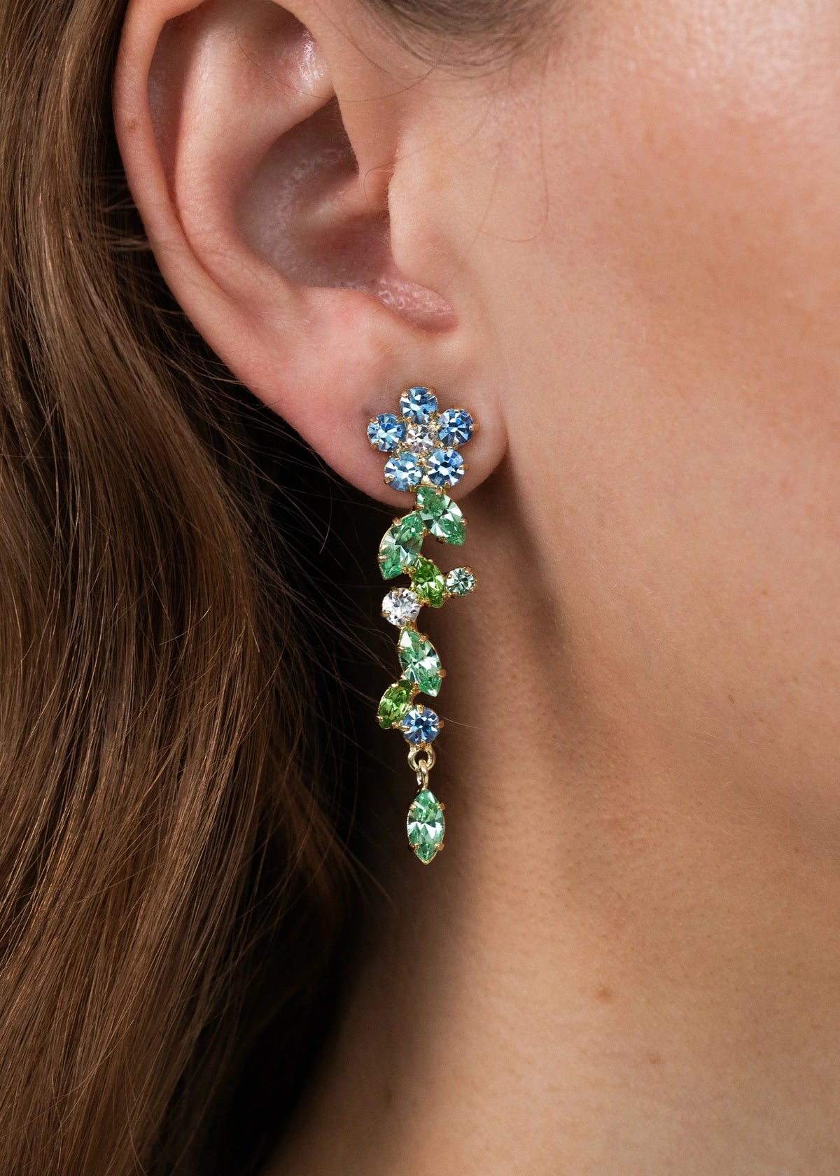 Close-up side view of Jasmina Earrings in Sky showing blue and green crystal floral cluster and dangling marquise stones in gold on a model’s ear