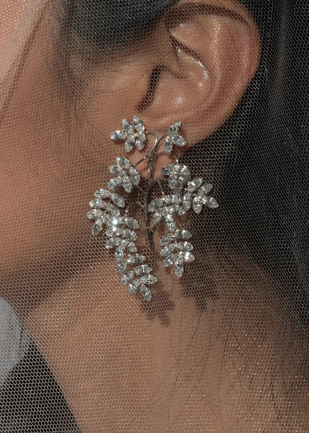 Close up of Raquel Earrings in Crystal showcasing vine-inspired cascading Austrian crystal clusters on a model’s ear behind a sheer veil