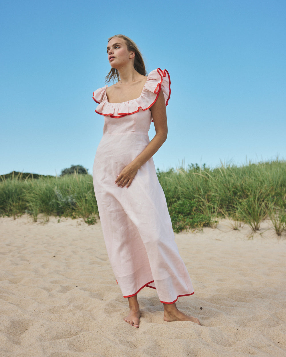 Full-length view of Parker Dress in pink sand on model with square neckline, ruffled red-trim sleeves, relaxed fit on beach