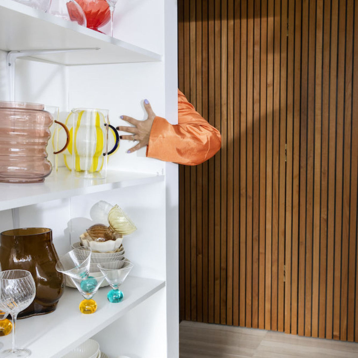 Yellow Curlie striped glass vase on white shelf among glassware, with a hand in an orange sleeve reaching from a wood wall