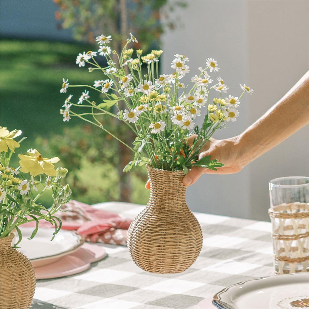 Hand placing a small basket weave resin vase filled with daisies on a checkered tablecloth with tableware in the background