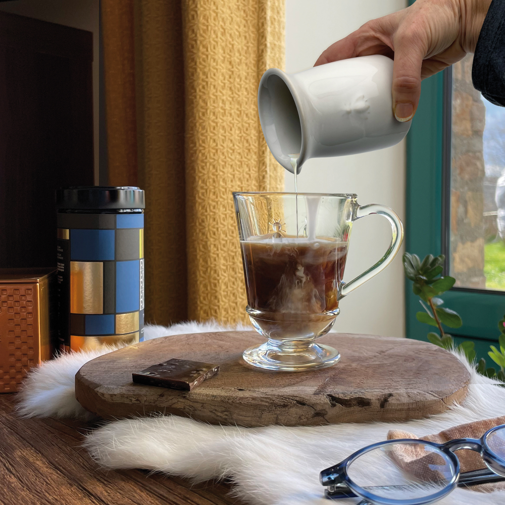 Ecru ceramic creamer pouring into glass coffee mug on wooden board with chocolate and reading glasses by a window