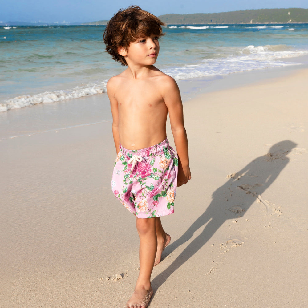 Three quarter view of Peony Boy Swim Trunk showing pink peony floral print and elastic drawstring waistband on a sandy beach