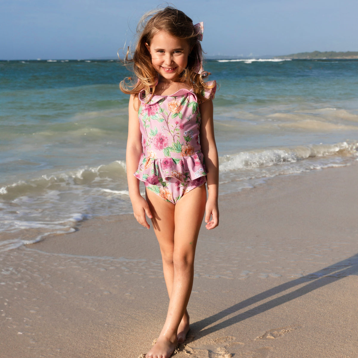 Front view of Peony Girl One-Piece showcasing pink floral print, ruffle neckline and tutu skirt with beach background
